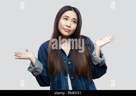 Non so. Confuso bella bruna asian giovane donna casual in denim blu con camicia con il trucco permanente i bracci sollevati e guardando lontano pensieroso. Foto Stock