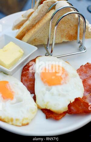 La prima colazione con uova fritte, pancetta, toast e rack di burro, il tutto servito su una piastra bianca Foto Stock