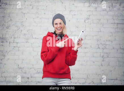 Piuttosto giovane donna sorridente in felpa rossa e Grey Hat con le cuffie sul suo collo in piedi vicino al muro di mattoni tenendo il telefono e dispositivo di puntamento su di esso. Foto Stock