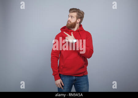 Bel giovane uomo barbuto che indossa felpa rossa in piedi su sfondo grigio e puntando il dito sul lato sinistro e guardando con il Foto Stock