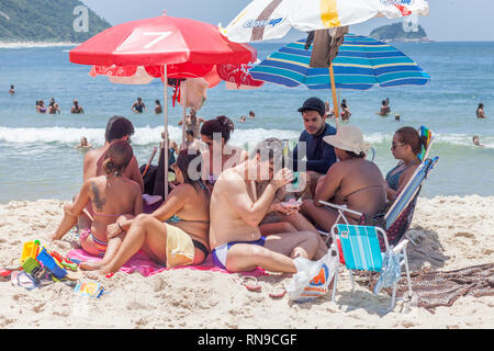 Frequentatori di spiaggia huddling insieme sotto all'ombra in una calda giornata estiva con persone in acqua in background Foto Stock