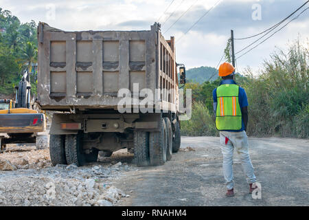 Costruzione maschio lavoratore di elmetto e giubbotto in piedi in strada per fermare il traffico veicolare durante il riposizionamento del carrello su strada sito in costruzione Foto Stock