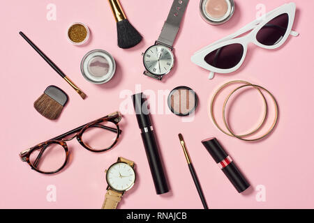 Vista dall'alto di orologi, rossetto, occhiali, occhiali da sole, ombretto, blush, spazzole cosmetiche, braccialetti e mascara su sfondo rosa Foto Stock