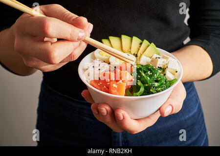 Mani femminili holding bacchette poke ciotola con il salmone, avocado riso, Chuka insalata, formaggio, uova di quaglia cosparso con il bianco e il nero lo sfondo di sesamo Foto Stock