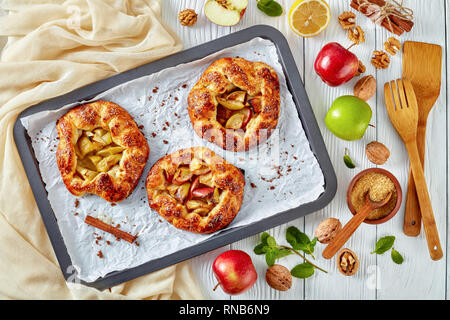 Aprire deliziose torte di mele, galette, con fette di mela ripieno su un vassoio da forno. apple, bastoncini di cannella, zucchero di canna e panno da cucina al backgrou Foto Stock