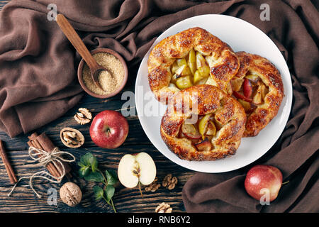 Aprire deliziose torte di mele, galette, con fette di mela su un piatto bianco. apple, bastoncini di cannella, zucchero di canna e panno da cucina su un vecchio tavolo rustico Foto Stock