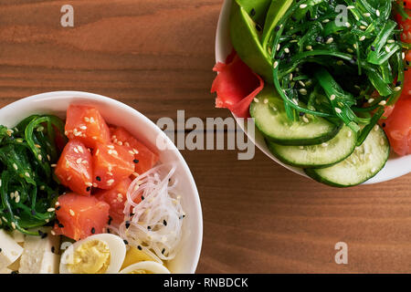 Materie organiche ciotola Poke con con riso, avocado, salmone, mango, cetrioli, chuka insalata di uova di quaglia dolce Cipolle due piastre di close-up su sfondo di legno Foto Stock