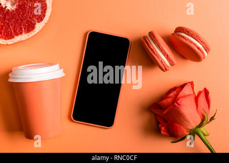 Smartphone in caso di corallo, coral bicchieri di carta, rose fiore, macarons e pompelmo metà su sfondo di corallo, il colore del concetto 2019 Foto Stock
