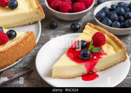 Cheesecake con la fetta di torta con lamponi freschi, mirtilli, marmellata e menta su calcestruzzo sfondo. close up. gustosa prima colazione Foto Stock