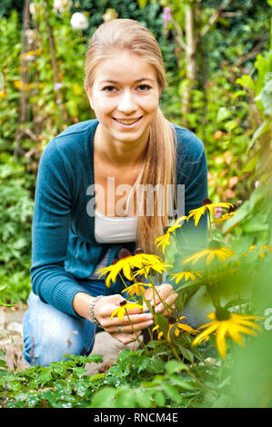 Junge Frau mit langen Haaren blondn hockt vor einem Strauch Sonnenhueten / Rudbeckia im Garten. [(C) Dirk A. Friedrich Foto Stock