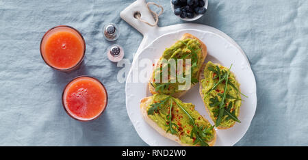 Banner di Avocado e sandwich di rucola con paprika sul tavolo. sana colazione o uno spuntino su una piastra su un lino blu tovaglia e spremere fresco Foto Stock