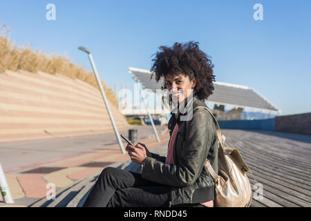 Donna nera con capelli afro seduti per strada con il suo smartphone in mano mentre sorride Foto Stock