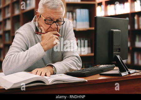 Senior uomo seduto in una classe e la lettura di un libro. Anziani uomo seduto in aula e di apprendimento con un libro e un computer sul tavolo. Foto Stock