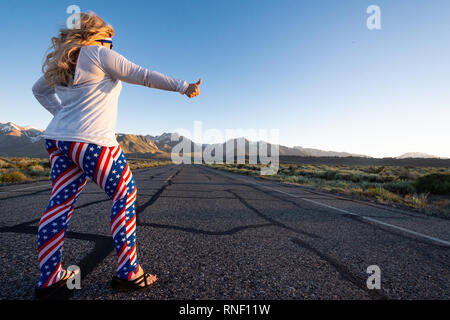 Femmina bionda indossando patriottica abbigliamento americano si erge nel mezzo di una strada di montagna con il suo pollice, concetto per autostop. Foto scattata in Foto Stock