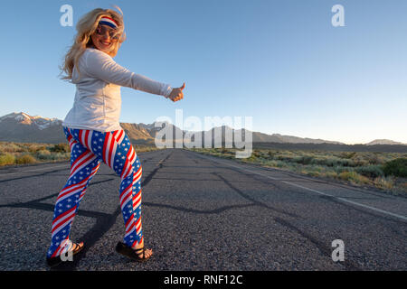 Femmina bionda indossando patriottica abbigliamento americano si erge nel mezzo di una strada di montagna con il suo pollice, concetto per autostop. Foto scattata in Foto Stock