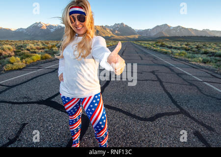 Femmina bionda indossando patriottica abbigliamento americano si erge nel mezzo di una strada di montagna con il suo pollice, concetto per autostop. Foto scattata in Foto Stock