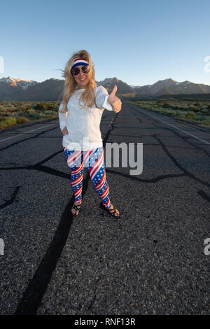 Femmina bionda indossando patriottica abbigliamento americano si erge nel mezzo di una strada di montagna con il suo pollice, concetto per autostop. Foto scattata in Foto Stock