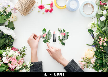 Vista ritagliata della mani femminili toccando rose boutonnieres vicino rossetto circondato da fiori su sfondo bianco Foto Stock