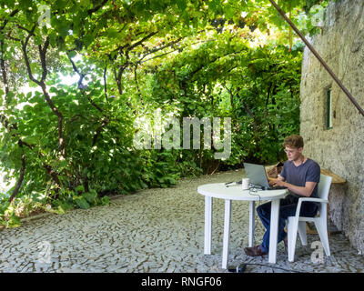 Un giovane uomo che lavora alla tastiera di computer portatile seduti sulla plastica sedia da giardino in fresco di ombra sotto grapevine vigneti di uve Foto Stock