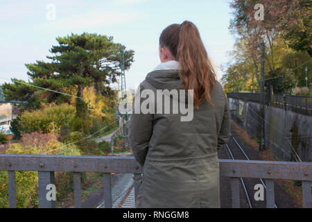 Ragazza adolescente è solo in piedi su un ponte e guarda i binari della ferrovia fotografato da dietro Foto Stock