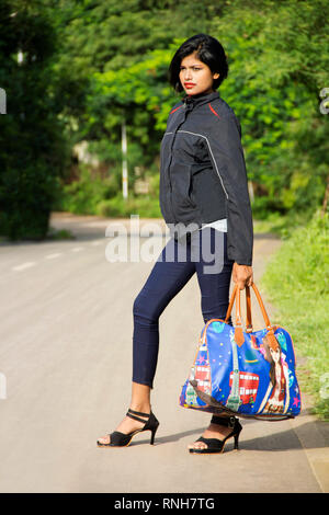 Giovane e bella ragazza indiana con peli corti holding di bagagli o borsa da viaggio in piedi a lato strada, Pune Foto Stock