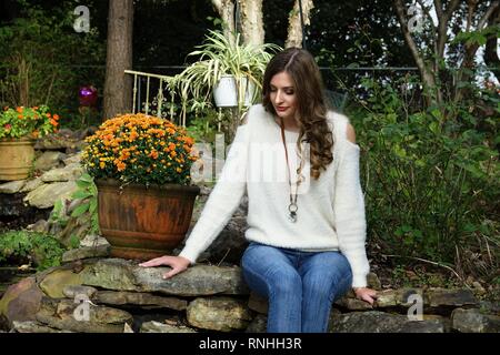 Paese giovane donna o ragazza con i capelli lunghi che indossa un maglione e jeans in un ambiente naturale con fiori e una parete di roccia Foto Stock