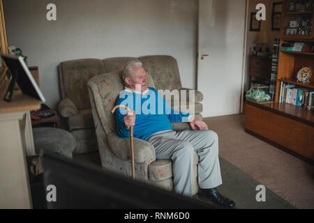L'uomo anziano è seduto nel suo soggiorno a casa con un bastone da passeggio. Foto Stock