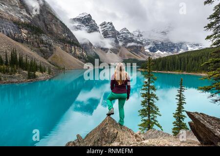 Giovane donna in piedi di fronte ad un lago cercando nel paesaggio di montagna, nuvole sospese tra picchi di montagna e la riflessione in turchese del Lago Moraine, La Foto Stock