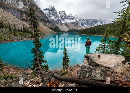 Giovane donna in piedi di fronte ad un lago cercando nel paesaggio di montagna, nuvole sospese tra picchi di montagna e la riflessione in turchese del Lago Moraine, La Foto Stock