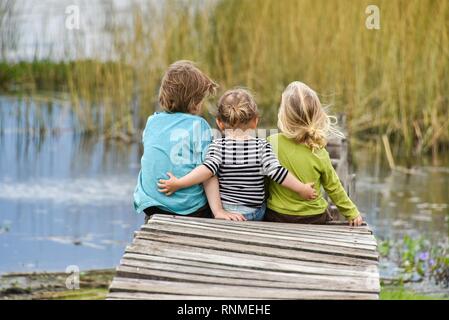 Tre bambini seduti insieme su un vecchio molo, vista posteriore, Patagonia, Argentina Foto Stock