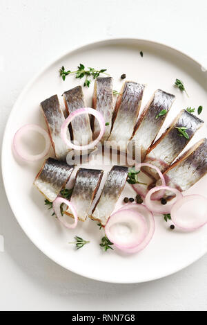Close up le aringhe salate di filetto con spezie e erbe aromatiche e la cipolla sulla piastra su pietra bianca dello sfondo. Fette di pesce marinato. Il cibo con una sana unsaturat Foto Stock