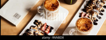 Banner dimensioni immagine di due tazze di latte caldo con cotto crosta di caramello e rotolo dolce con banana e fragola sul tavolo di legno in coffee-shop. Caffè Foto Stock