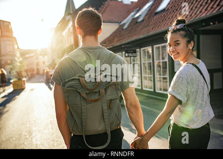 Sorridente giovane donna guardando sopra la sua spalla mentre camminando mano nella mano con il suo fidanzato su una strada di città nel pomeriggio Foto Stock