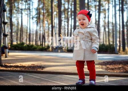 Bionda dolce piccolo bimbo ragazza nel bellissimo beige trench coat e cappuccio rosso svolge all'aperto presso il parco giochi Foto Stock