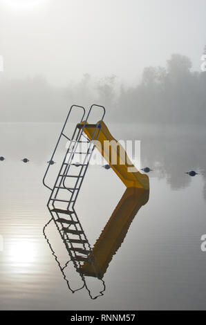 La riflessione di una diapositiva giallo in una nebbiosa mattina da un fiume Foto Stock