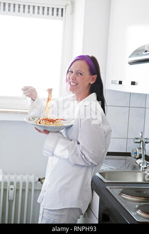 Junge weiss gekleidete Frau lehnt in ihrer Kueche an der Arbeitsplatte und haelt einen Teller Spegetti in der mano. Sie hat eine violett gefaerbte str Foto Stock