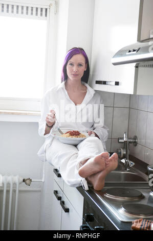 Junge weiss gekleidete Frau sitzt in ihrer Kueche auf der Arbeitsplatte und haelt einen Teller mit spaghetti in der mano. Sie hat eine violett gefaer Foto Stock