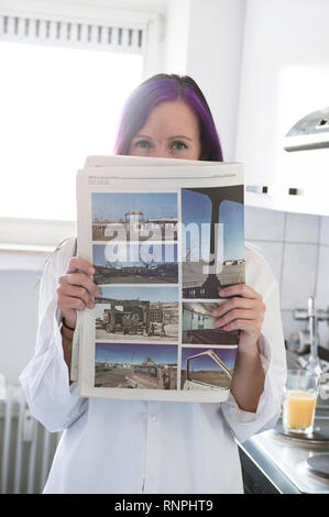 Junge weiss gekleidete Frau steht in ihrer Kueche an der Arbeitsplatte und haelt eine Zeitung in der mano. Sie hat eine violett gefaerbte Straehne im Foto Stock