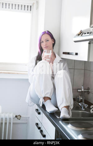 Junge weiss gekleidete Frau sitzt in ihrer Kueche auf der Arbeitsplatte und haelt Ein Glas Milch in der mano. Sie hat eine violett gefaerbte Straehne Foto Stock