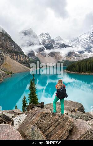 Giovane donna in piedi di fronte ad un lago cercando nel paesaggio di montagna, nuvole sospese tra picchi di montagna e la riflessione in turchese del Lago Moraine, La Foto Stock