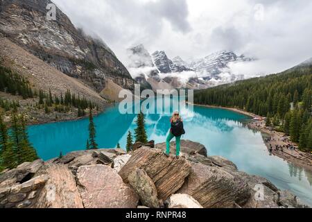 Giovane donna in piedi di fronte ad un lago cercando nel paesaggio di montagna, nuvole sospese tra picchi di montagna e la riflessione in turchese del Lago Moraine, La Foto Stock