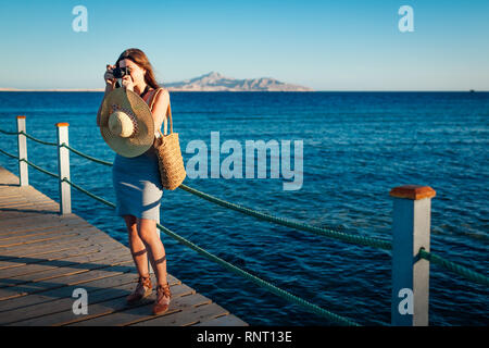 Giovane donna traveler per scattare delle foto del paesaggio di mare sul molo l'utilizzo della fotocamera. Moda estate. La vacanza estiva. Foto Stock