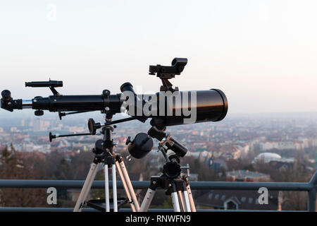 Due telescopi professionali, uno newtoniani e un rifrattore, montati su treppiedi su una terrazza sul tetto, guardando una grande città al tramonto. Foto Stock