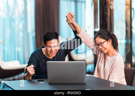 Giovani asiatici coppia sposata tenere mani celebrando insieme, guardando il computer portatile in modern home office. Lavoro di successo, amore concetto di relazione Foto Stock