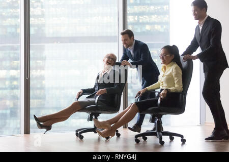 Felice diversi dipendenti divertirsi a cavallo su sedie in ufficio Foto Stock