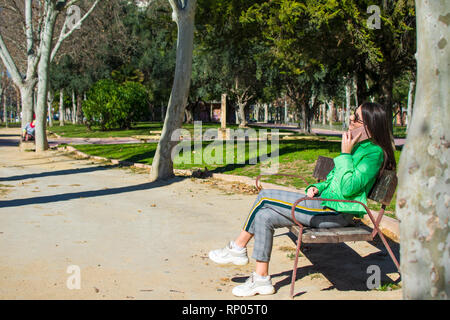 Felice lonely donna o ragazza seduto su una panchina nel parco sorridente in autunno o caduta stagione in attesa di qualcuno e utilizzando il suo telefono, Murcia, Spagna, 2019. Foto Stock