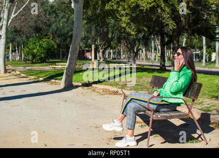 Felice lonely donna o ragazza seduto su una panchina nel parco sorridente in autunno o caduta stagione in attesa di qualcuno e utilizzando il suo telefono, Murcia, Spagna, 2019. Foto Stock