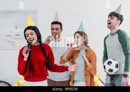 Amici multiculturale in colorate per i cappelli di carta a cantare il karaoke a casa party Foto Stock