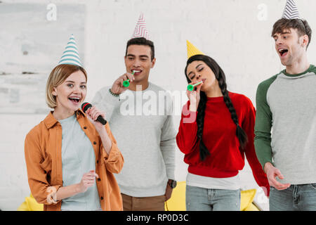 Multietnica amici multiculturale in colorate per i cappelli di carta avendo divertimento a casa parte cantando il karaoke Foto Stock