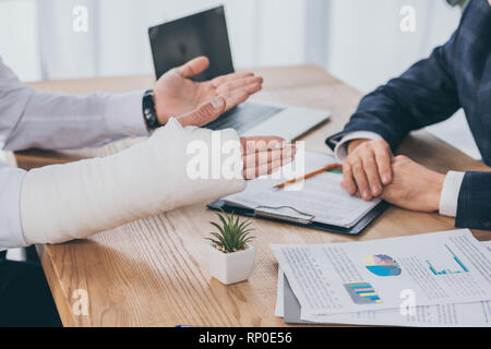 Vista ritagliata del lavoratore con braccio rotto ubicazione a tavola con i documenti di fronte imprenditore in ufficio, concetto di compensazione Foto Stock